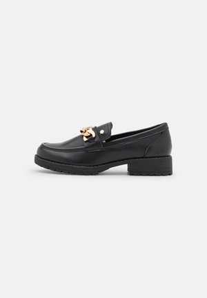 Mocassin en cuir noir avec un petit talon bloc et un détail de chaîne en or sur le dessus, présenté de profil sur un fond blanc.