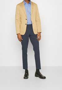 Béžové sako na dvě knoflíky přes světle modrou košili, doplněné tmavě modrými kalhotami a černými mokasínami s texturovanou tkaninou.