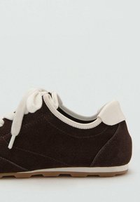 Zapatilla de ante marrón con un acento en el talón color crema, cordones blancos y una suela de goma texturizada, con un diseño redondeado y de perfil bajo.