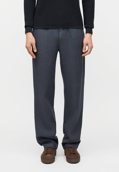 Homme portant un pantalon droit gris foncé, des chaussures marron à lacets et un haut à manches longues noir, debout devant un fond uni.