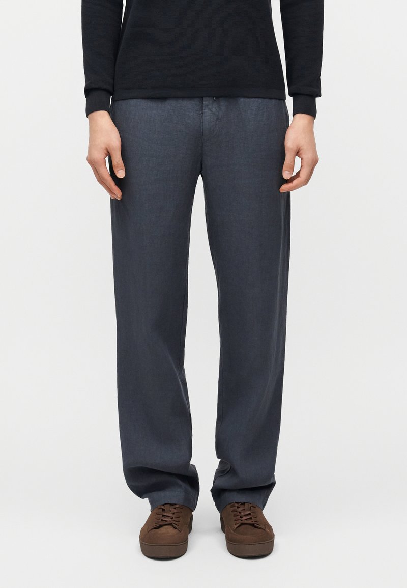 Άνδρας που φοράει σκούρα γκρι παντελόνια με ίσια πόδια, καφέ δερμάτινα παπούτσια με κορδόνια και μαύρο μπλουζάκι μακριάς μανίκας, στέκεται μπροστά σε απλό φόντο.