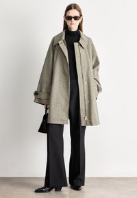 Manteau à motif pied-de-poule en beige et noir, doté d'un grand col, de fermetures à boutons, et d'une silhouette décontractée. Associé à un pantalon noir et des chaussures noires.