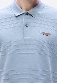 Γαλάζιο πόλο πουκάμισο με υφή στο ύφασμα, με οριζόντιες ρίγες και πατιλέτα με τρία κουμπιά. Διαθέτει καφέ μεταλλικό λογότυπο ως διακοσμητικό στοιχείο.