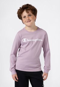 Chemise lavande à manches longues avec un logo "Champion" blanc, en tissu doux, associée à un pantalon de jogging noir avec des accents de logo.