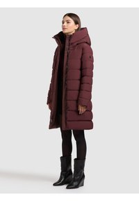 Bordový prešívaný kabát s vysokým golierom, zapínaním na gombíky a bočnými vreckami, doplnený čiernymi členkovými topánkami, na neutrálnom pozadí.