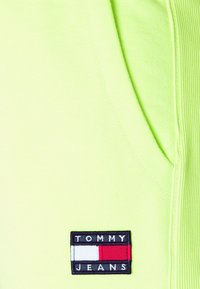 Neon zelená látka s hladkým povrchem a boční kapsou. Obsahuje obdélníkovou námořnickou etiketou s nápisem "TOMMY JEANS" v bílé a červené barvě.