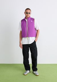 Un jeune homme portant un gilet sans manches violet, un T-shirt blanc, un pantalon cargo noir, des baskets argentées et des lunettes de soleil, se tenant sur un sol vert contre un mur blanc.