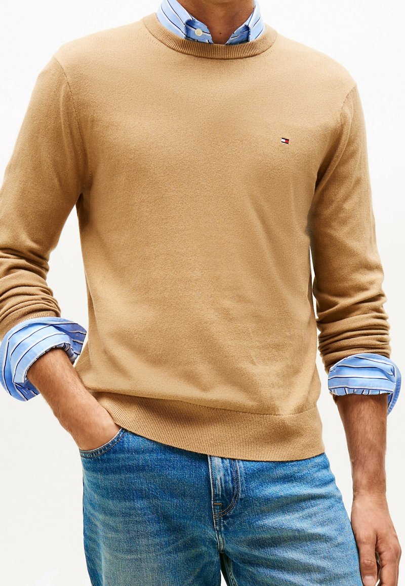Homme portant un pull beige par-dessus une chemise rayée bleue avec les manches retroussées, associé à un jean bleu, main dans la poche.