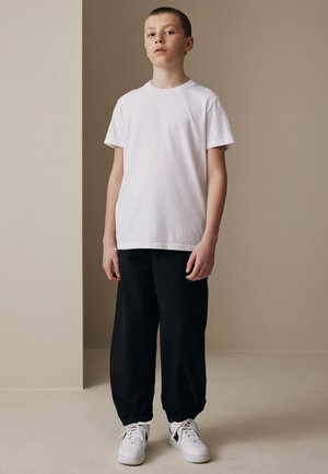 Niño de pie en el interior con una camiseta blanca lisa, pantalones negros sueltos y zapatillas blancas sobre un fondo beige.