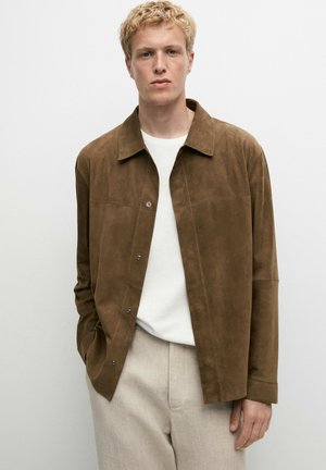 Junger Mann mit blondem Haar trägt eine braune Wildlederjacke über einem weißen Hemd und hellbeigen Hosen vor einem schlichten weißen Hintergrund.