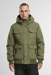 Chaqueta acolchada verde oliva con capucha, cuatro bolsillos frontales y puños elásticos. Diseñada con cierre de cremallera y dobladillo acanalado.