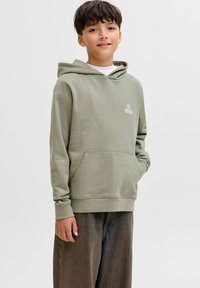 Chlapec s krátkymi tmavými vlasmi, oblečený v svetlozelenom mikinovom kapucni a tmavých nohaviciach, stojí s jednou rukou v vrecku na jednoduchom pozadí.