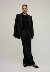 Blazer preto ajustado com botões duplos, combinado com calças pretas de cintura alta. Sapatos de salto agudo pretos completam o conjunto.