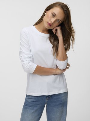 Jeune femme aux longs cheveux bruns portant une chemise blanche à manches longues et un jean bleu, reposant sa tête sur sa main.
