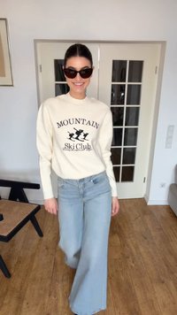 Γυναίκα με γυαλιά ηλίου, πουλόβερ κρεμ με το κείμενο "Mountain Ski Club", ανοιχτό μπλε τζιν με φαρδιά μπατζάκια, στέκεται σε δωμάτιο με ξύλινο πάτωμα και γυάλινες πόρτες.