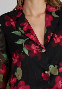Blusa negra con estampado floral que presenta flores rojas y hojas verdes, fabricada en tejido transparente, con cuello con muesca y detalles de botones en tono dorado.
