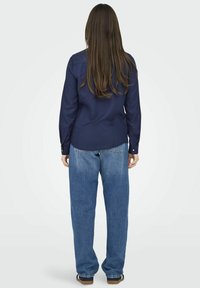 Camicia blu navy con bottoni e maniche lunghe, abbinata a jeans blu a vestibilità rilassata. Il tessuto sembra liscio; la camicia ha un yoke posteriore per una migliore vestibilità.