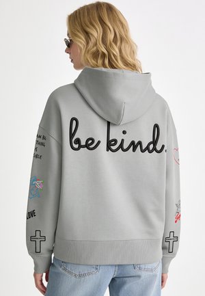 Donna con capelli biondi che indossa una felpa con cappuccio grigio chiaro con testo "be kind." sulla schiena e varie piccole grafiche sulle maniche, di spalle.