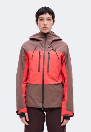 Personne portant une veste à capuche marron et rouge avec plusieurs poches verticales zippées, debout devant un fond clair uni.