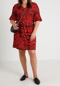 Mujer vistiendo un vestido de estampado de tigre rojo y negro, sosteniendo un bolso cruzado negro, con zapatos negros de estilo deslizante sobre un fondo plano.