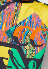 Chemise à manches courtes jaune vif et vert, présentant un motif abstrait multicolore vibrant et des détails graphiques noirs audacieux.