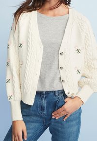 Cárdigan de punto trenzado crema con bordado floral en rosa y verde, con botones plateados, que se lleva sobre una camiseta gris y jeans azules.