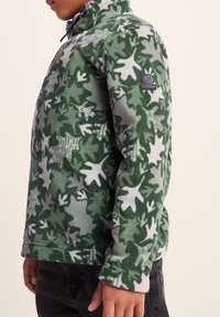 Green fleece jacket with a leaf pattern in varying shades, featuring a high collar and logo patch on the left sleeve.