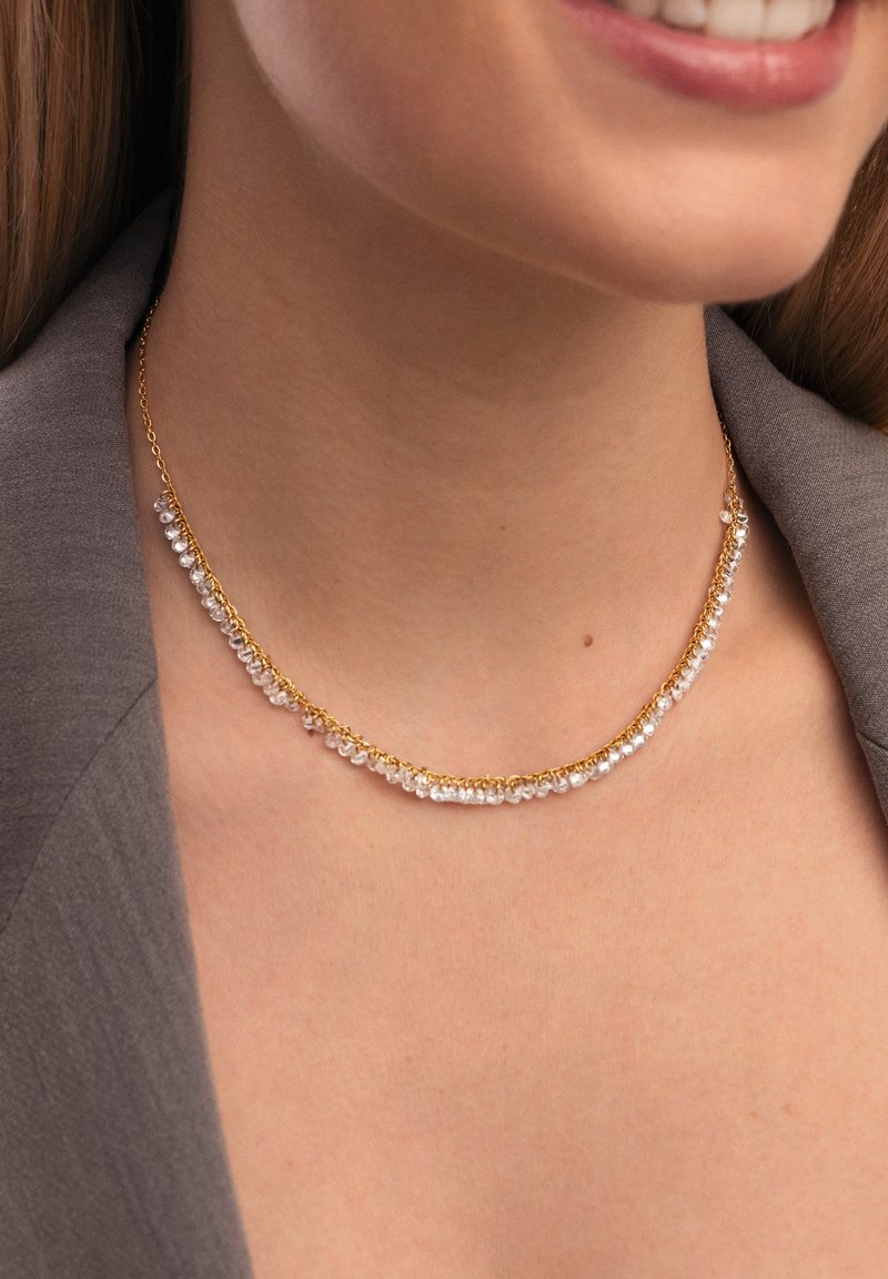 Collana in oro con perline di cristallo trasparente, disposte in una linea curvata, che evidenzia una superficie liscia e riflettente con una sottile texture.