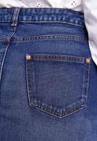 Jeans de mezclilla azul oscuro con un gran bolsillo trasero que incluye un remache de cobre, acentuado por costuras contrastantes. Textura suave y ajuste clásico.