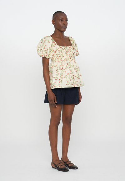 Ivory-kukkakuosisella peplum-puserolla, jossa on lyhyet puff-hihat, yhdistetty tummansinisiin shortseihin ja tummiin tasapohjaisiin kenkiin. Puserossa on neliönmallinen pääntie.