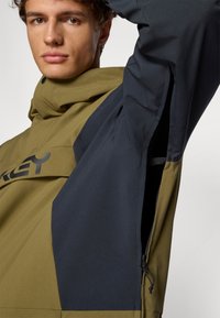 Giacca verde e blu con cappuccio, dotata di una grande tasca frontale e dettagli con zip laterale. Realizzata in tessuto liscio e resistente all'acqua.