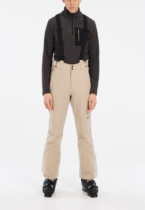 Beige skibroek met verstelbare zwarte bretels, ritssluiting en een relaxte pasvorm. Draagt met een donker langlopend shirt en zwarte skischoenen.
