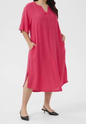 Donna che indossa un vestito rosa acceso fino al ginocchio, con maniche corte, spacchi laterali, tasche e scarpe slingback nere con tacco basso.