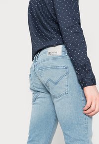 Person som bär ljusblå denimjeans med en bakficketikett och en marinblå långärmad skjorta med ett litet vitt mönster.