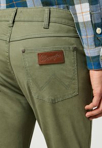 Pantalones Wrangler de color verde oliva con un parche de cuero en el bolsillo trasero, costuras distintivas y una tela de textura suave.
