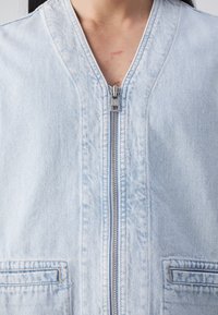 Světlomodrá džínová bunda se zipem vpředu, stojáčkovým límcem, dvěma předními kapsami a prošívanými detaily podél švů.