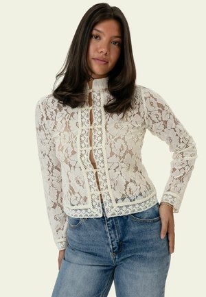 Kvinde med langt mørkt hår iført en hvid floral blondebluse med lange ærmer og blå jeans, poserer med den ene hånd på hoften.