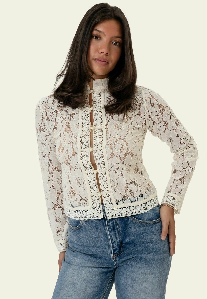 Femme aux longs cheveux foncés portant un top en dentelle blanche à fleurs à manches longues et un jean bleu, posant avec une main sur la hanche.