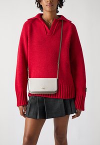 Maglione rosso lavorato a maglia con colletto, abbinato a una gonna plissettata a quadri neri. Viene indossata una piccola borsa a tracolla color crema con dettagli dorati.
