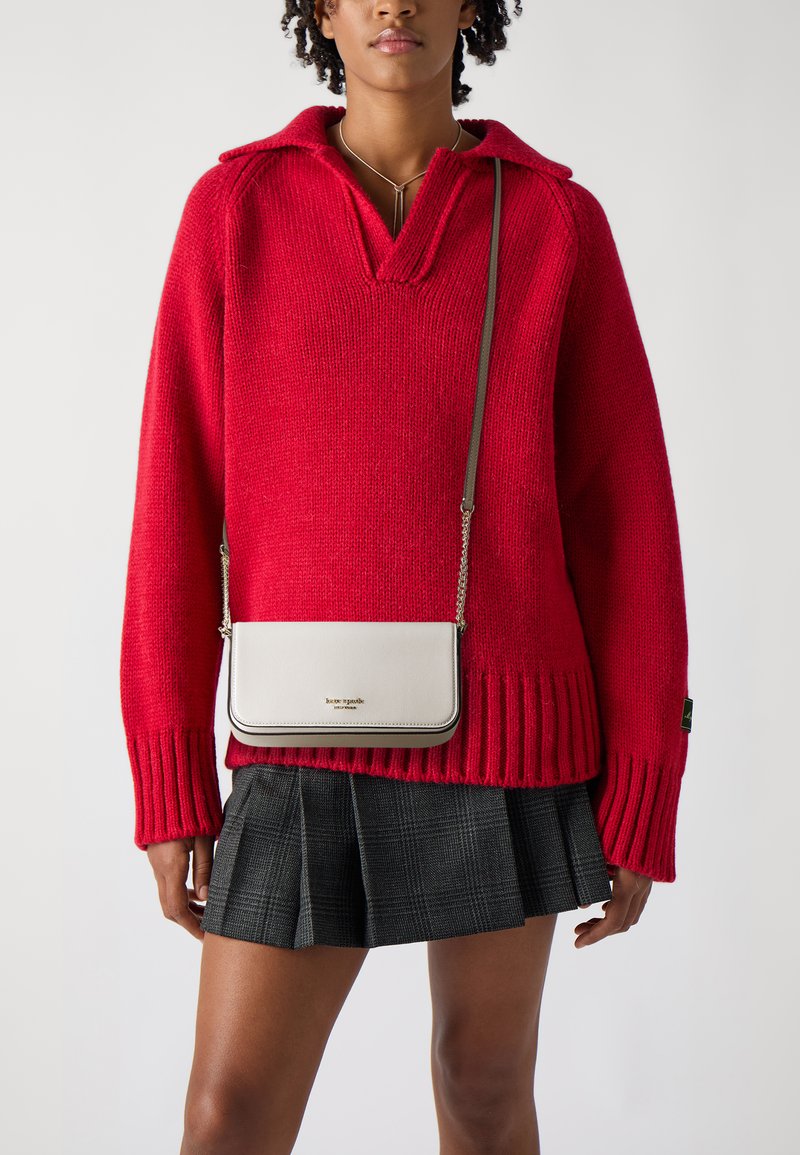 Maglione rosso lavorato a maglia con colletto, abbinato a una gonna plissettata a quadri neri. Viene indossata una piccola borsa a tracolla color crema con dettagli dorati.