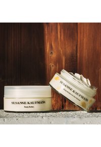 Des contenants de beurre corporel blanc avec des couvercles transparents. L'un est scellé, et l'autre montre une texture crémeuse. Les étiquettes indiquent "Susanne Kaufmann."
