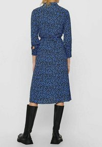 Abito blu con stampa leopardata nera, polsini abbottonati e cintura in vita. Di lunghezza media, realizzato in un tessuto liscio. Abbinato a stivali neri fino al ginocchio.