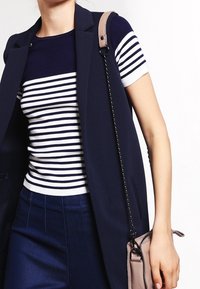 Chaleco sin mangas azul marino sobre una camiseta de rayas azul marino y blanca, combinado con pantalones azules, persona sosteniendo un pequeño bolso con cadena.