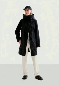 Cappotto impermeabile nero con collo alto, chiusura frontale a scatto e tasche laterali, abbinato a un cappello nero, una camicia fantasia e pantaloni chiari.