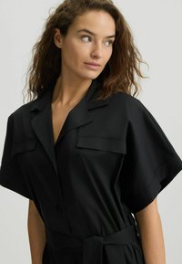 Abito nero con bottoni, maniche corte, due tasche sul petto, cintura in vita e texture liscia. Design semplice e silhouette classica.