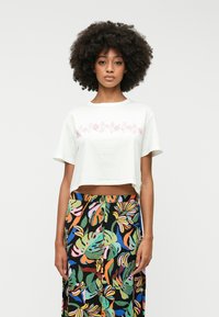 White cropped T-shirt with pink floral print, paired with a long black skirt featuring colorful leaf patterns and button details.