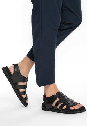 Füße in schwarzen, ledernen Riemchensandalen, kombiniert mit dunkelblauen, strukturierten Hosen vor weißem Hintergrund.