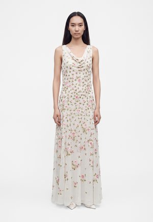 Femme aux longs cheveux foncés portant une robe blanche longue sans manches avec une broderie florale rose et verte, debout sur un fond blanc.