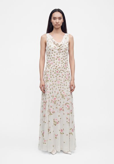 Femme aux longs cheveux foncés portant une robe blanche longue sans manches avec une broderie florale rose et verte, debout sur un fond blanc.