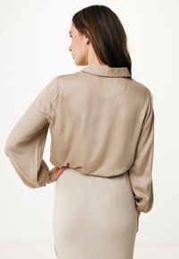 Béžová saténová košile s dlouhými, nadýchanými rukávy a klasickým límcem, v kombinaci se světle řasenou sukní. Hladká textura, minimalistický design.